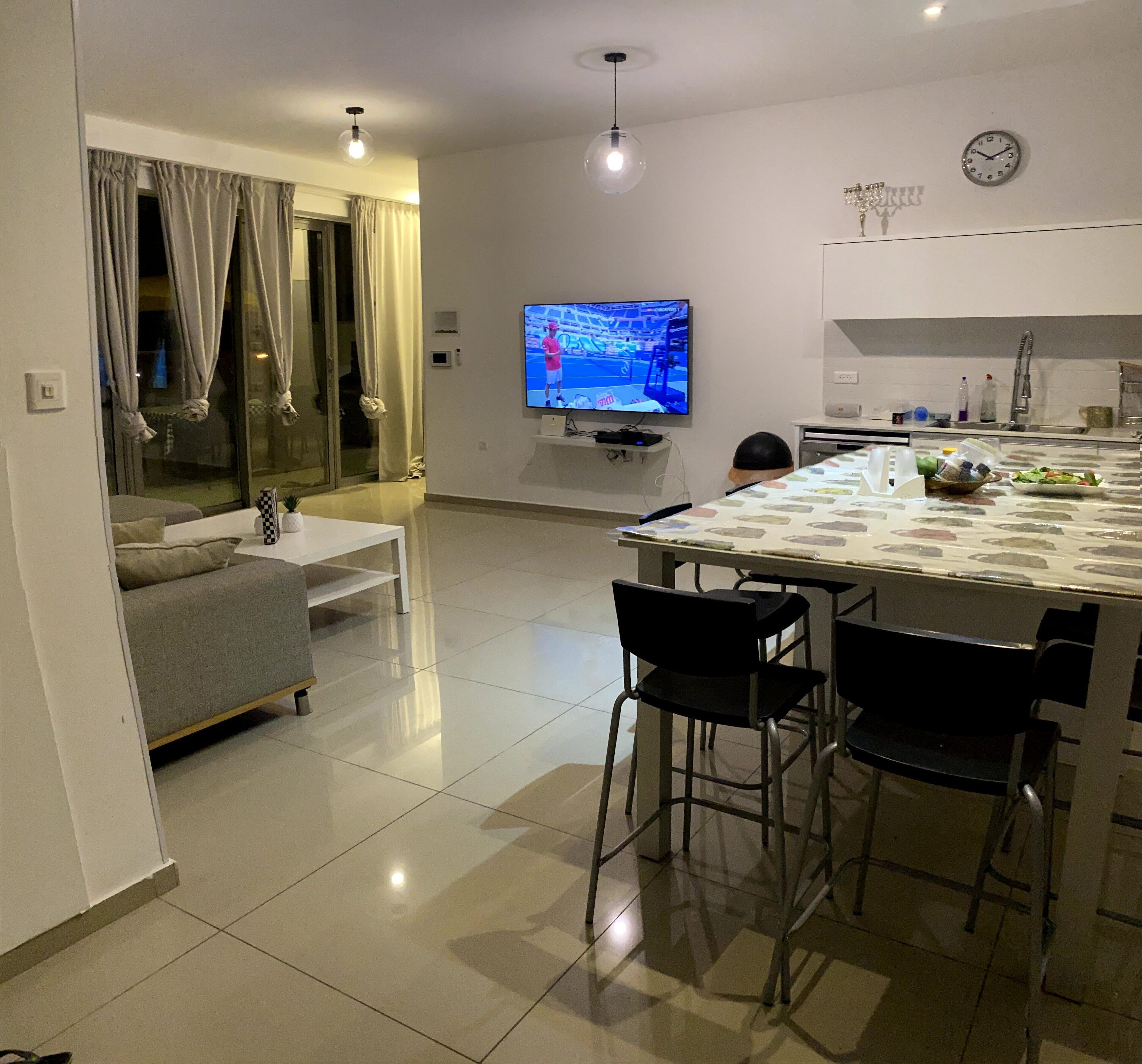 A modern living room with a flat-screen TV, a dining area with a table and chairs, and a kitchen area with a sink and countertop.