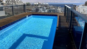 Une piscine sur le toit, avec une eau d’un bleu limpide, entourée d’une terrasse en bois et de rampes de protection en verre. En arrière-plan, on aperçoit des bâtiments et un paysage montagneux.
