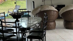 Een eethoek met een glazen tafel en stoelen, een aan de muur bevestigde televisie, rieten stoelen en uitzicht op de omgeving.