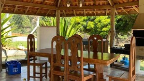 Een overdekte buitenruimte met een houten tafel en stoelen, een plafondventilator en een barbecue op de achtergrond.