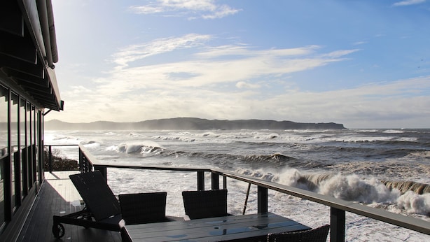 Une terrasse avec deux chaises et une table, d’où l’on peut admirer une mer agitée et les vagues qui se brisent sur le rivage.