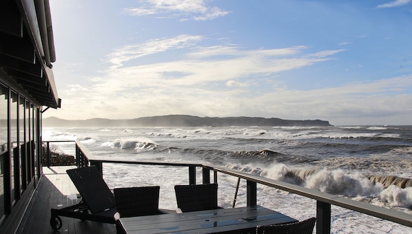 A deck with two chairs and a table overlooking a turbulent sea with waves crashing onto the shore.