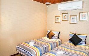 A room with two beds, each with striped bedding and pillows, a wall-mounted air conditioner, and framed pictures on the wall.