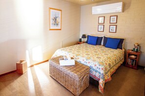 A bedroom with a bed, a nightstand, a lamp, a small table with a book, a wicker basket, and a wall with framed pictures. - 'Seabreeze' a unique Waterfront/Beachside property (Pearl Beach)