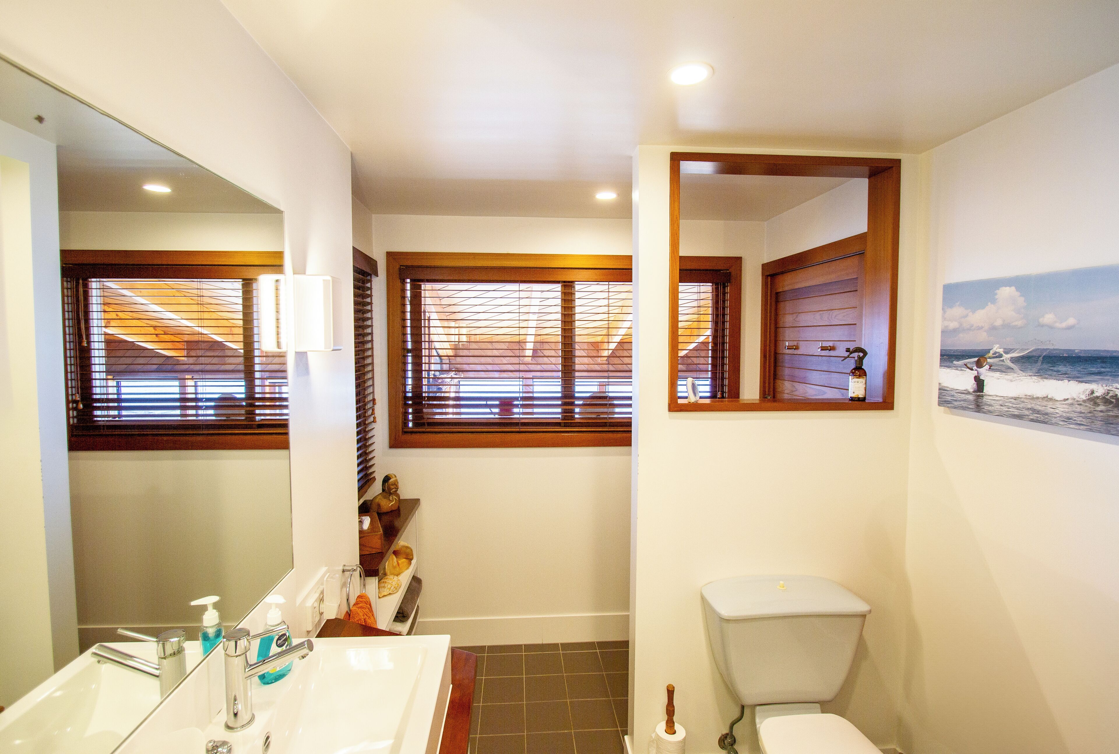 A bathroom with a large mirror, a sink with faucets, a toilet, and a window with blinds.