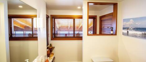 A bathroom with a large mirror, a sink with faucets, a toilet, and a window with blinds.