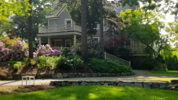 A two-story house with a balcony, surrounded by lush greenery and flowering plants. - Harvard House--Make Your 2026 Family Gathering An Unforgettable Experience (Waltham)