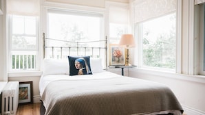 A bedroom with a bed, a nightstand, a lamp, and a window with a view of trees. - Harvard House--Make Your 2026 Family Gathering An Unforgettable Experience (Waltham)