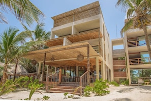 A multi-story building with a covered outdoor area, surrounded by palm trees and a sandy ground. - Hotel Los Arrecifes (Tulum)