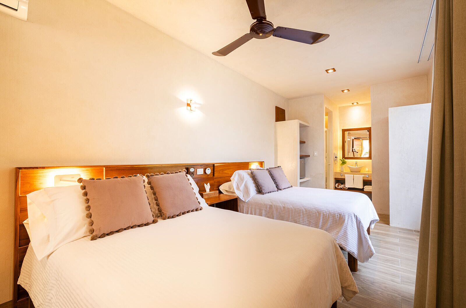Habitación de hotel con dos camas, ventilador de techo, mesita de noche con lámpara y baño visible a través de una puerta abierta.