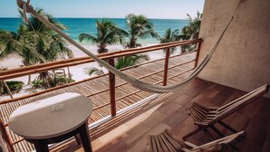 A balcony with a hammock, wooden chairs, and a table overlooking a beach with palm trees and the ocean. - Hotel Los Arrecifes (Tulum)