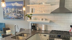 Una cocina moderna con estufa de acero inoxidable, un lavaplatos y una cafetera. Hay un cuadro de paisaje urbano en la pared y una planta en maceta sobre la encimera.