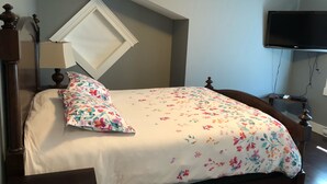 Un dormitorio con cama, mesita de noche, lámpara, televisor y ventana con persiana blanca.