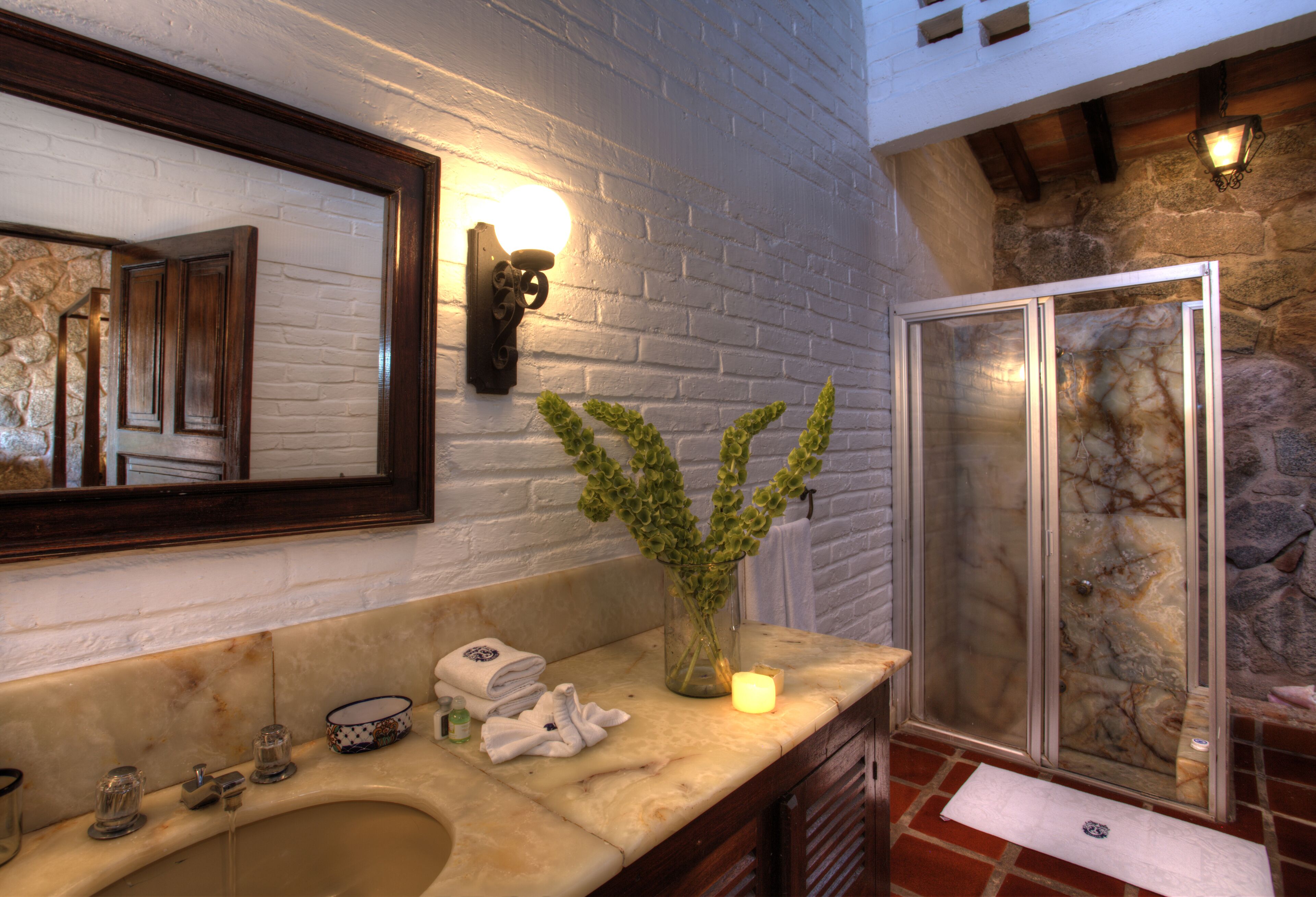 A bathroom with a marble sink, a mirror, a vase with green plants, a lit candle, and a glass shower enclosure.