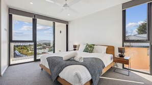 Un dormitorio moderno con una cama grande, una mesita de noche, una lámpara y vistas a un paisaje urbano a través de amplios ventanales.