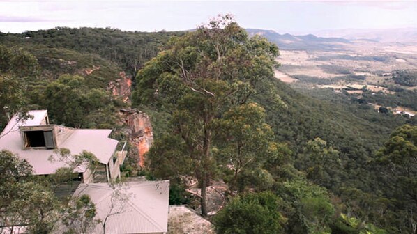 Une maison avec un toit en pente et une cheminée bien visible, entourée d’une forêt dense et de collines.