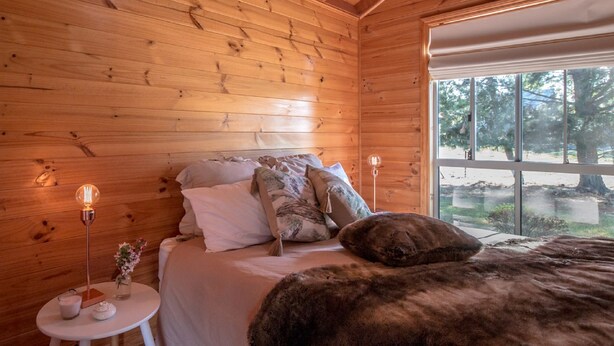 Une chambre à coucher dans une cabane en bois, avec un lit, des oreillers, une table de chevet avec une lampe et une fenêtre donnant sur les arbres.