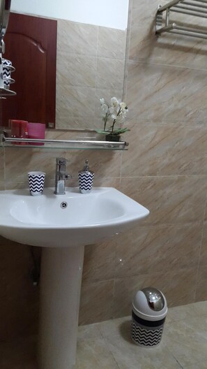 Ein Badezimmer mit einem weißen Waschbecken, zwei gemusterten Tassen, einer kleinen Topforchidee und einem Mülleimer.