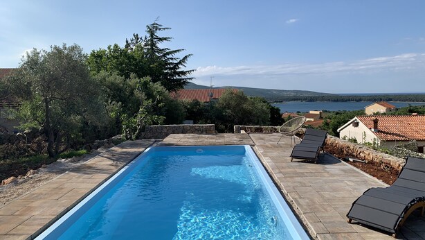 Una piscina rectangular amb aigua blava i cristal·lina, envoltada de rajoles de pedra. Hi ha gandules i una paret de pedra. Al fons, hi ha arbres, un edifici amb teulada vermella i una vista d'una massa d'aigua i les muntanyes.