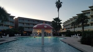 A swimming pool with a mushroom-shaped water feature, surrounded by lounge chairs and palm trees, with hotel buildings in the background. - Beachfront condo near Top Places! Heated pool in Winter (Galveston)