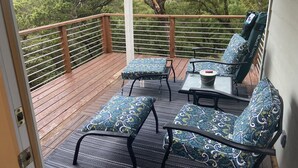A balcony with a wooden deck, two patterned chairs, a small table, and a view of a forested hillside.