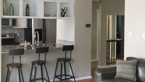 A modern kitchen with a central island, bar stools, and a dining area with a sofa and a patterned pillow.