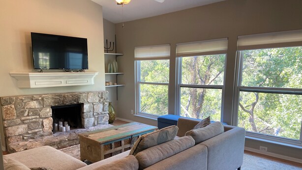 A living room with a stone fireplace, a flat-screen TV, and large windows with a view of trees.