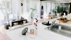 A modern living room with a large window, a white sofa, a coffee table, and a kitchen area with a sink and a wooden bowl.