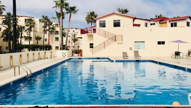 A swimming pool with a staircase leading to a building, surrounded by palm trees and a clear sky. - BEAUTIFUL CONDO IN ROSARITO (VILLA SERENA) (Predios Urbanos)