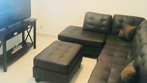 A living room with a sectional sofa, a TV on a stand, and a small ottoman. - BEAUTIFUL CONDO IN ROSARITO (VILLA SERENA) (Predios Urbanos)