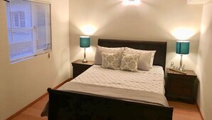 A neatly arranged bedroom with a bed, two bedside tables, a wall-mounted light, and a window with blinds. - BEAUTIFUL CONDO IN ROSARITO (VILLA SERENA) (Predios Urbanos)