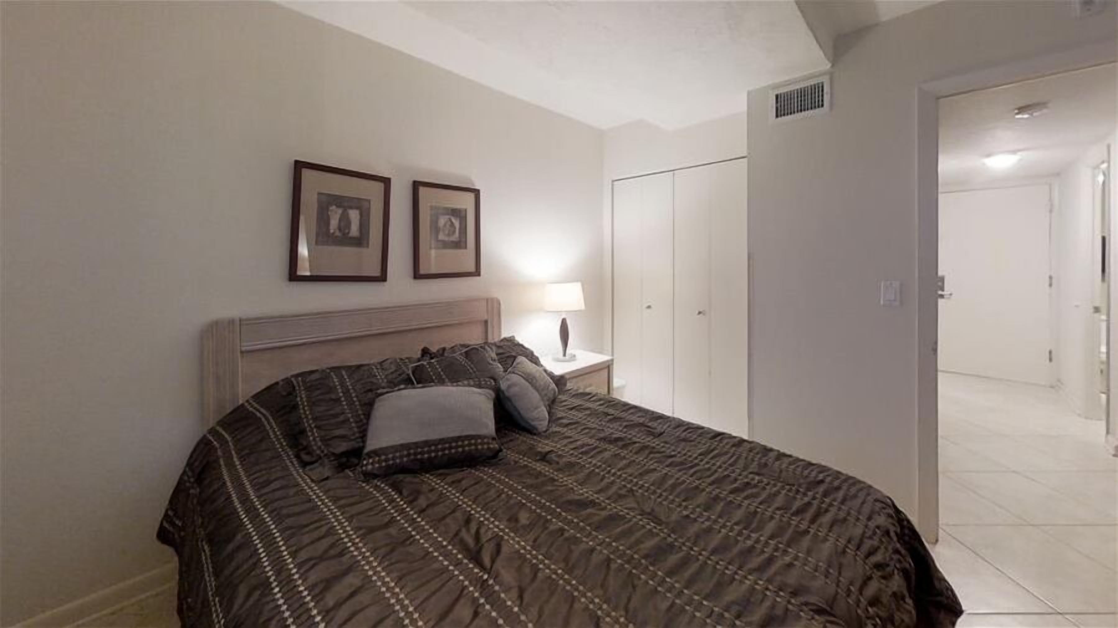 A bedroom with a bed, a nightstand with a lamp, two framed pictures on the wall, and a wardrobe.