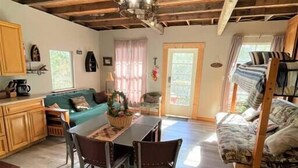 A rustic cabin interior with a dining area, wooden ceiling beams, a chandelier, a sofa, and a bunk bed. - The Cedar House at Hamilton Creek (Burnet County)
