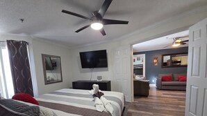 Une chambre à coucher comprenant un lit, un ventilateur de plafond, une télévision, une commode et une vue sur un espace de vie avec un canapé et un coin repas.