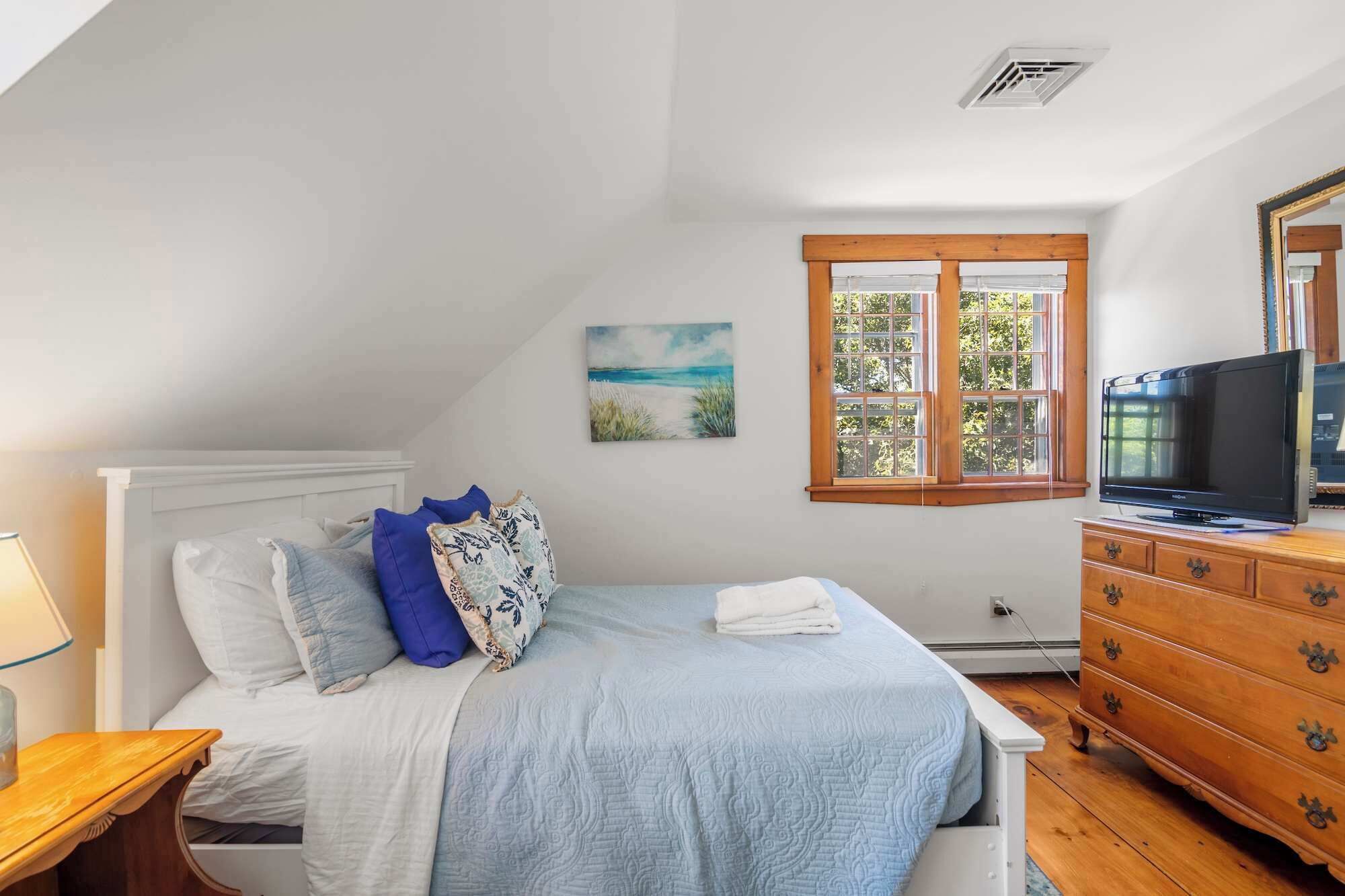 A bedroom with a bed, a dresser, a television, and a window with a view of greenery.