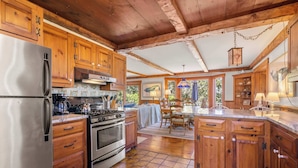 A spacious kitchen with wooden cabinets, a stainless steel refrigerator, a stove, and a dining area with a table and chairs. - Stunning Water Views 2 Decks! By Beach! (Chatham)