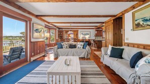 A spacious living room with a large window overlooking a beach, wooden beams on the ceiling, and a cozy seating area with striped cushions. - Stunning Water Views 2 Decks! By Beach! (Chatham)
