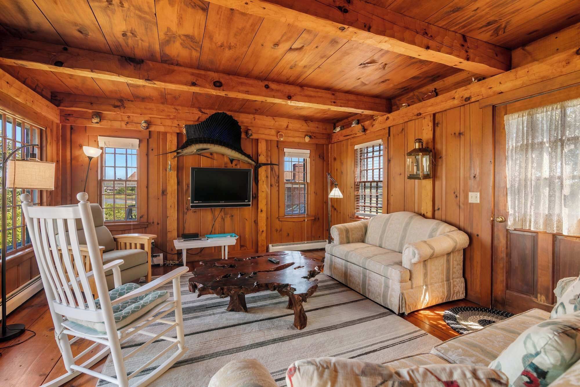 A cozy cabin interior with wooden walls, a mounted television, a rocking chair, a sofa, and a unique wooden coffee table.