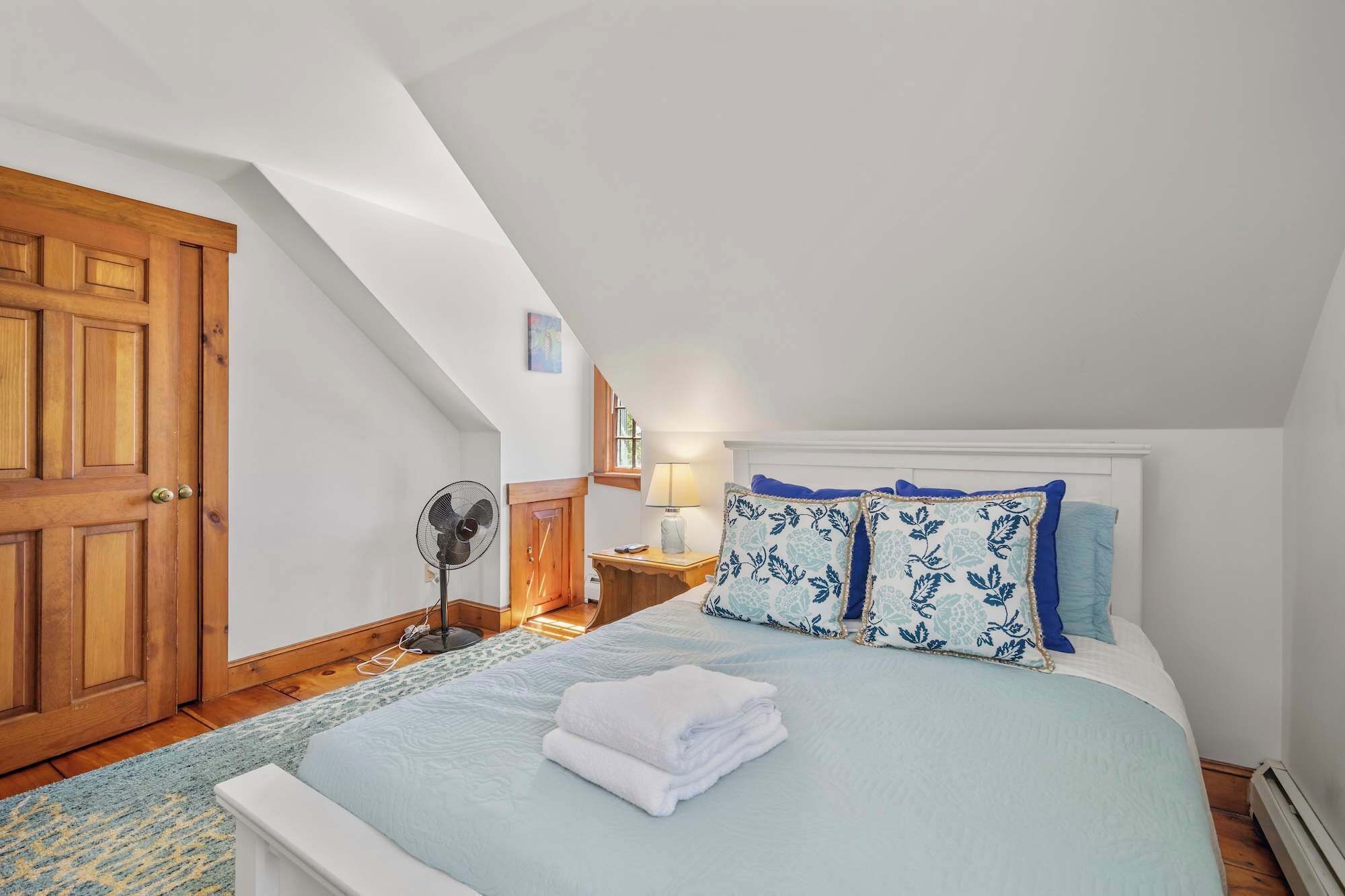 Un dormitorio con techo inclinado, una puerta de madera, una cama con almohadas de diseño azul y blanco, una lámpara de noche y un ventilador.