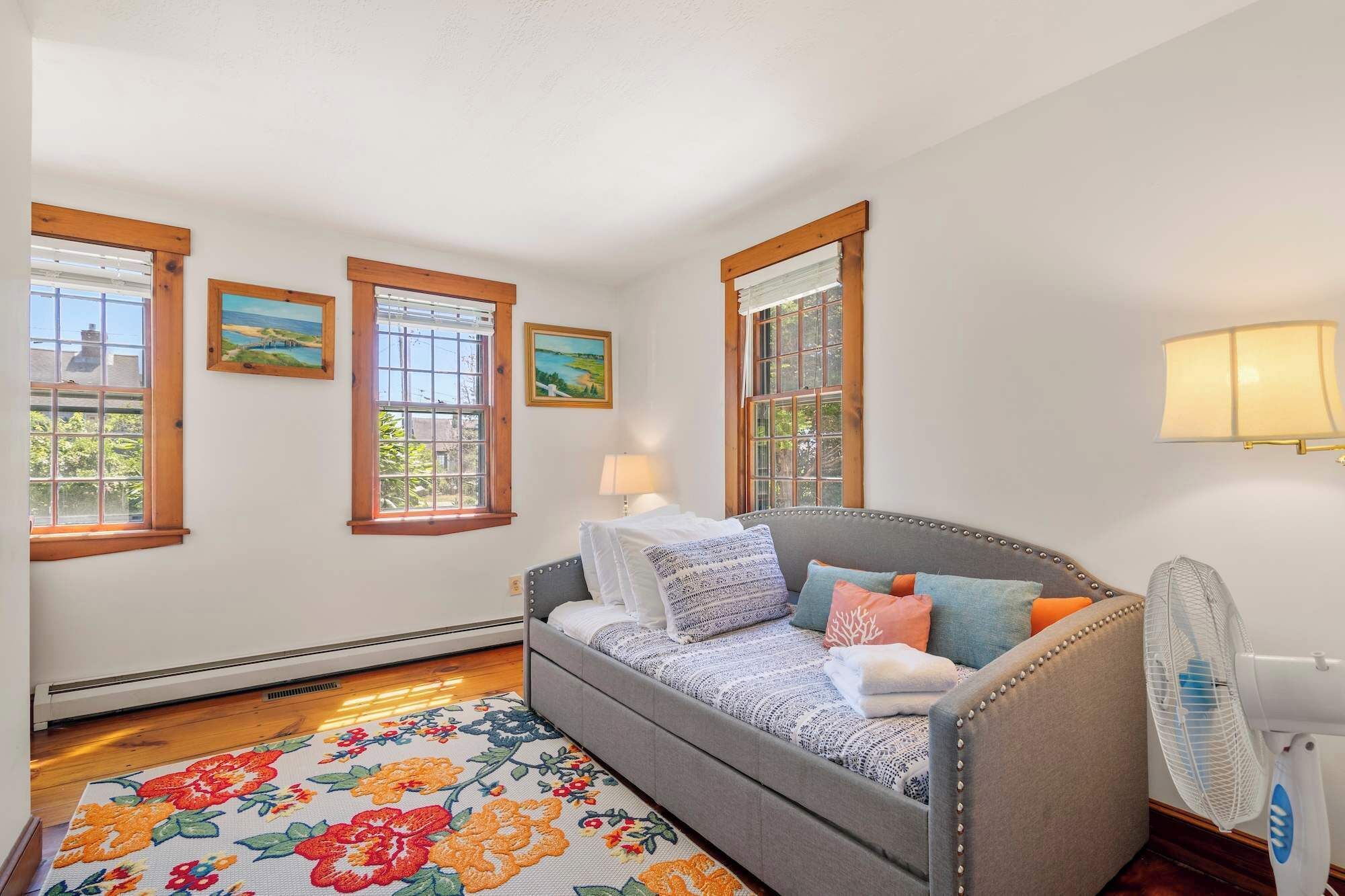 Una habitación acogedora con un sofá gris, alfombra floral y tres ventanas con marcos de madera.