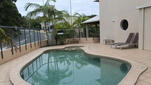 Une piscine à bord incurvé, entourée d’une terrasse carrelée et d’une clôture en verre. On y trouve des chaises longues et un banc près de la piscine.