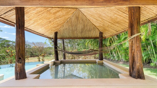 A pool area with a thatched roof, surrounded by lush greenery.