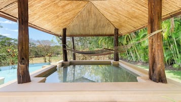 A pool area with a thatched roof, surrounded by lush greenery.