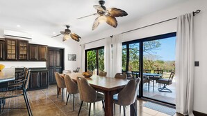 A dining area with a large table and chairs, a kitchen with dark cabinets, and a ceiling fan.