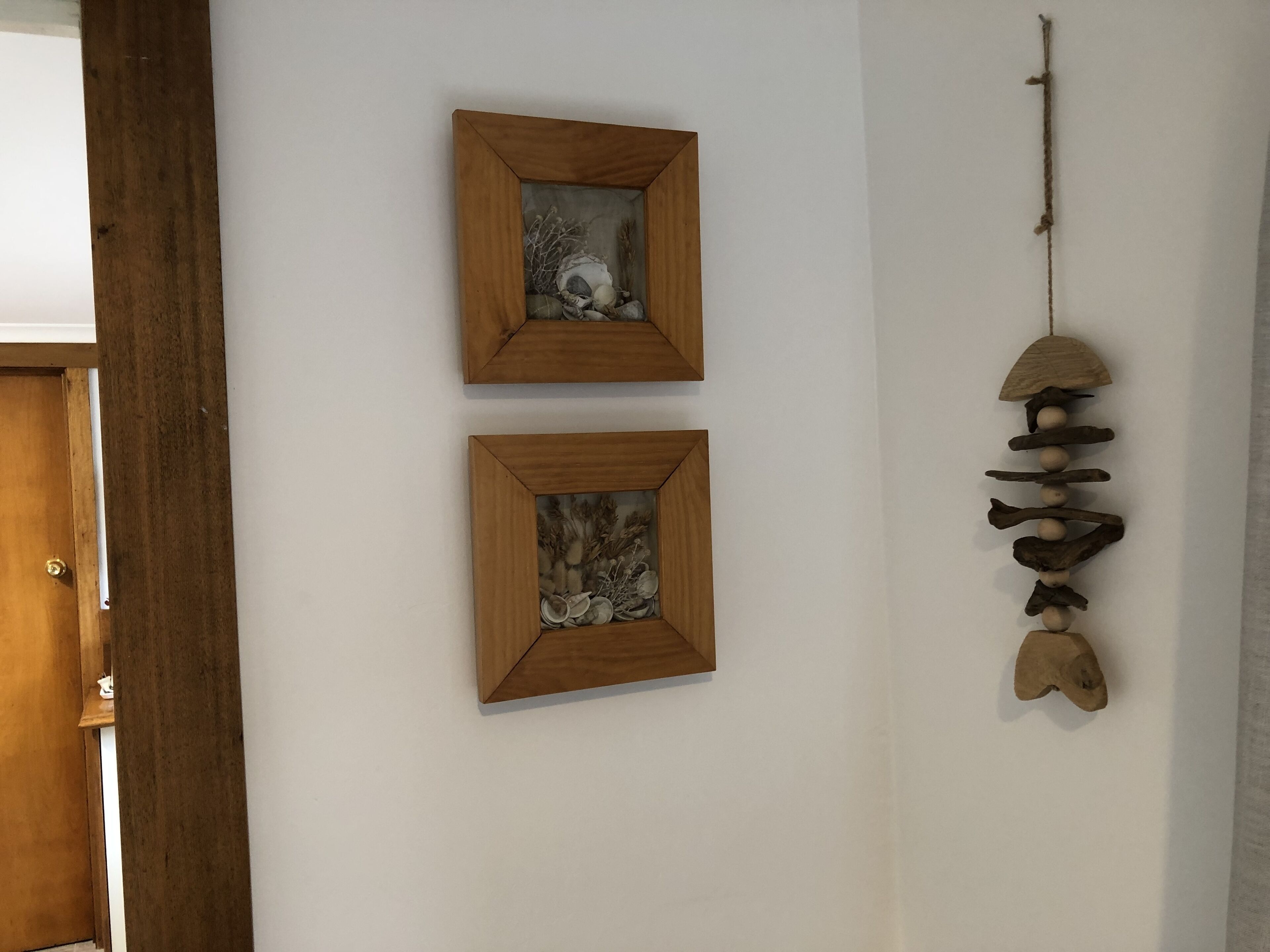 Two framed artworks on a white wall, a wooden door to the left, and a hanging wind chime to the right.