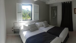 A bedroom with a bed, two bedside tables, a window with a view of greenery, and a door. - CASA DA VIÑA (Nogueira de Ramuín - Ourense) Ribeira Sacra  (Ourense)
