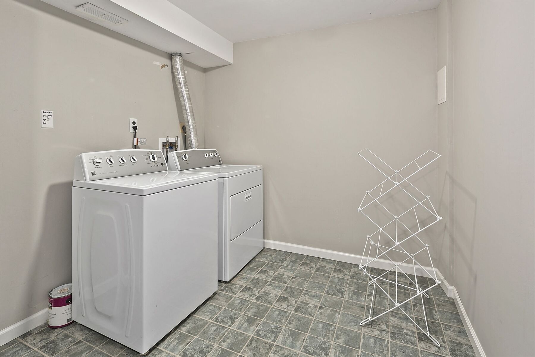A laundry room with two front-loading washing machines, a drying rack, and a vent above the machines.