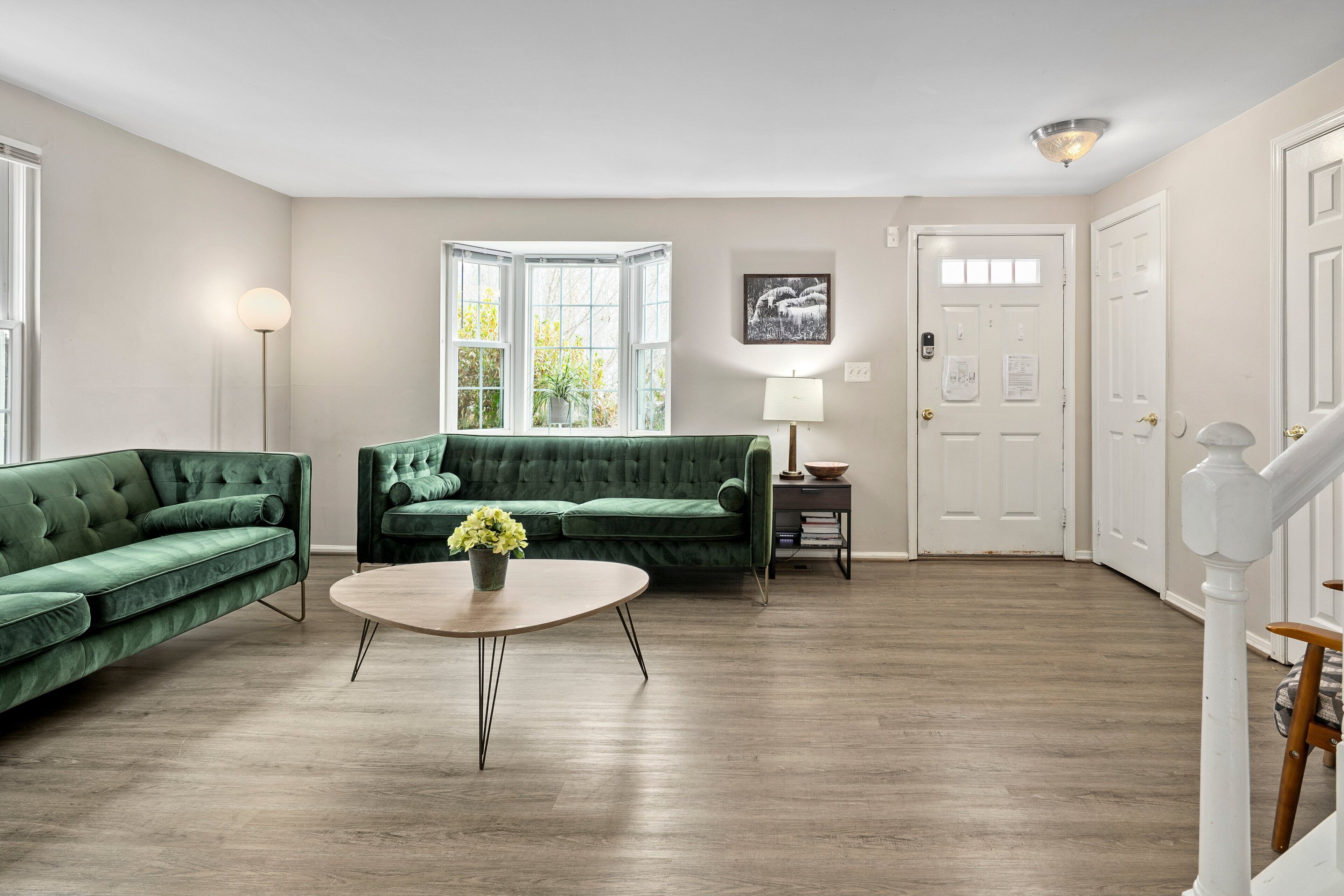 Een ruime woonkamer met een groene bank, een ronde salontafel met bloemen, een lamp en uitzicht door een groot raam.