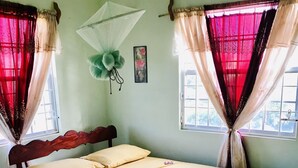 A bed with a blue bedspread and a white mattress, a wooden headboard, and a window with red and white curtains. - YOUR HOME AWAY FROM HOME (Vieux Fort)
