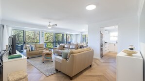 Ein großzügiger Wohnbereich mit einer großen Couch, einer kleineren Couch und einem Couchtisch. Es gibt einen Essbereich mit Tisch und Stühlen. Der Raum verfügt über große Fenster und einen Deckenventilator.